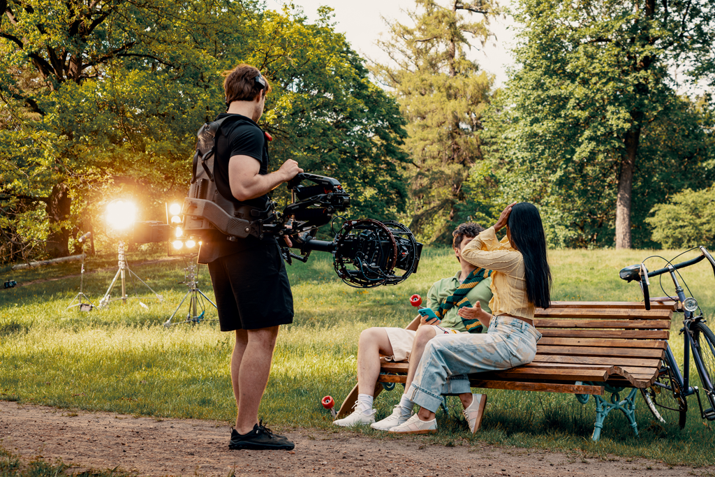 Operator z kamerą podczas zdjęć plenerowych do filmu reklamowego kręconego we Wrocławiu, scena z aktorami na ławce w parku