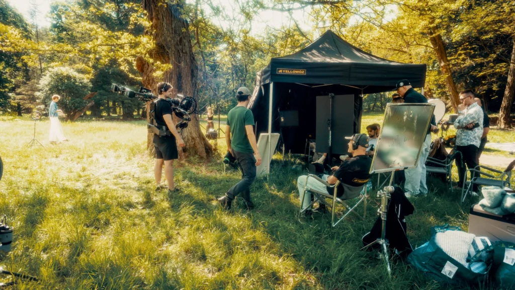 Produkcja filmowa – plan zdjęciowy spotu reklamowego Chillberry w parku: operator z kamerą, stanowisko podglądu i ekipa filmowa